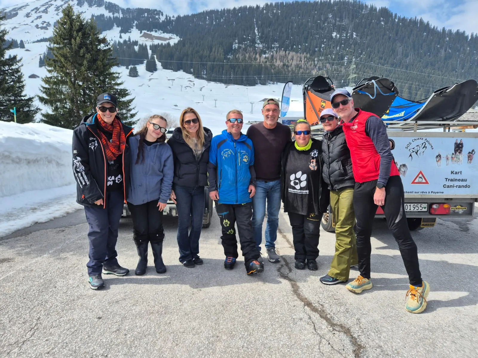 Huit personnes réunies sur une route de montagne enneigée, devant la remorque de l’association marquée « SOS Chiens Polaires — kart, traîneau, cani-rando »