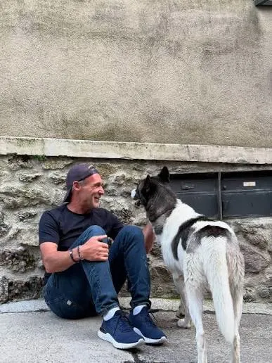 Mike Horn, assis au sol contre un mur, souriant à un chien noir et blanc debout à côté de lui